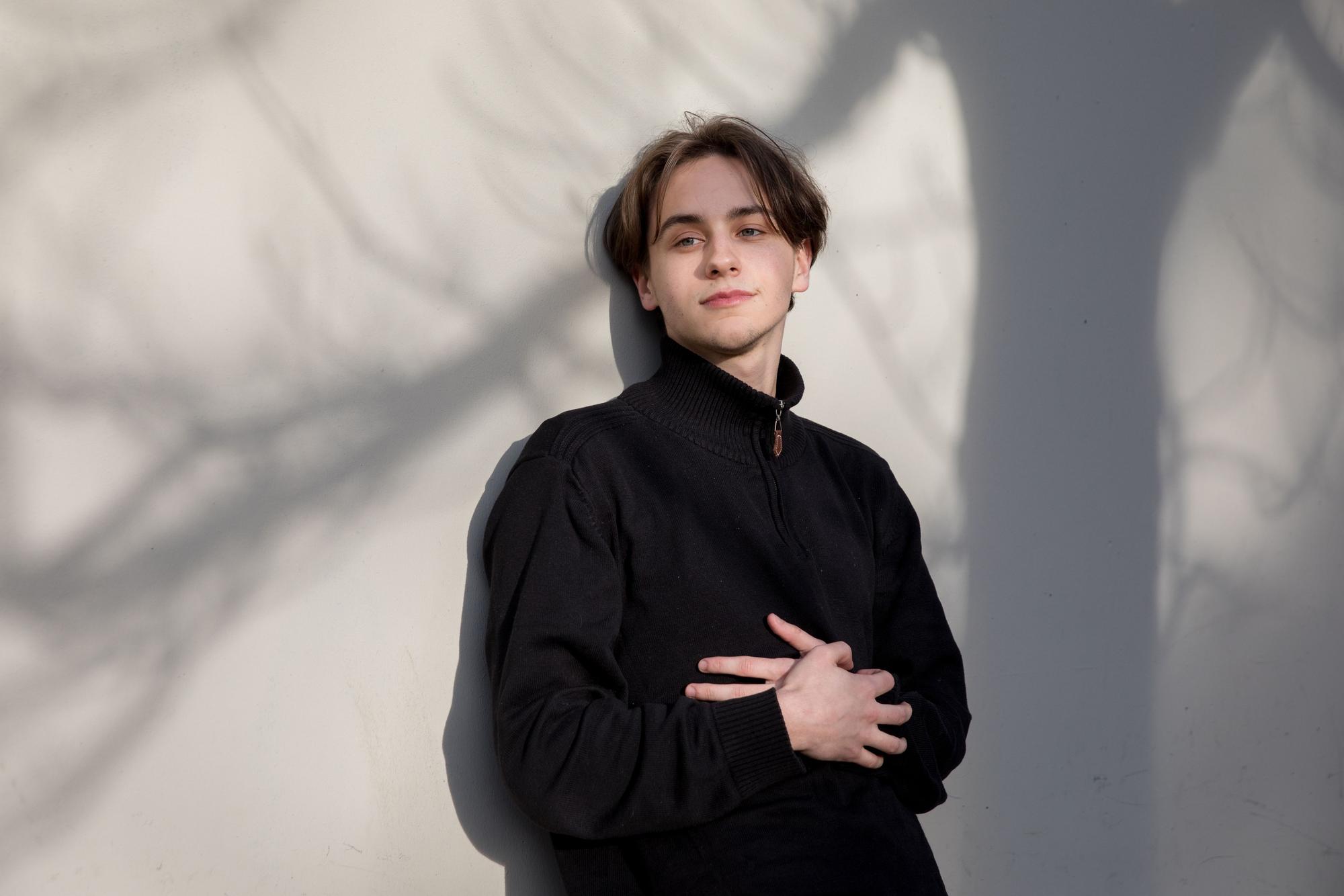 Tristan Simon lehnt an einer weißen Wand, auf der die Schatten eines Baumes zu sehen sind.
