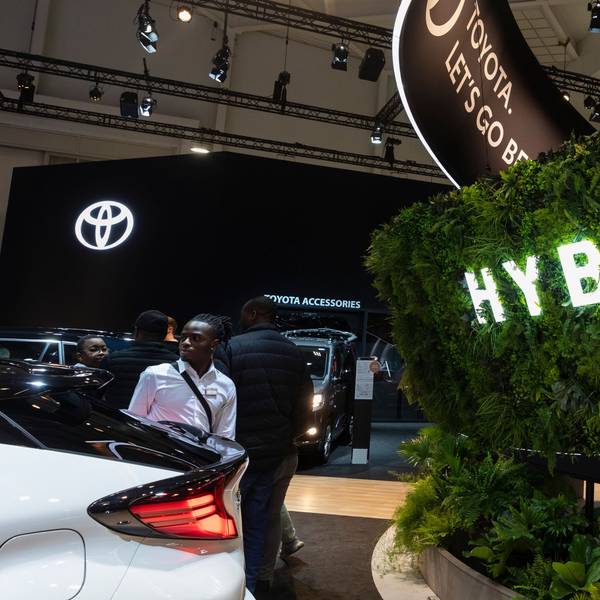 Abgebildet ist der Brüsseler Autosalon. Es sind Menschen und Autos sowie das Toyota-Logo zu sehen. Im Vordergrund ist eine Hecke mit einer Leuchtaufschrift „Hybrid“ zu sehen.