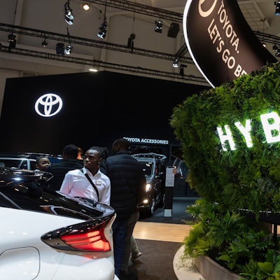 Abgebildet ist der Brüsseler Autosalon. Es sind Menschen und Autos sowie das Toyota-Logo zu sehen. Im Vordergrund ist eine Hecke mit einer Leuchtaufschrift „Hybrid“ zu sehen.