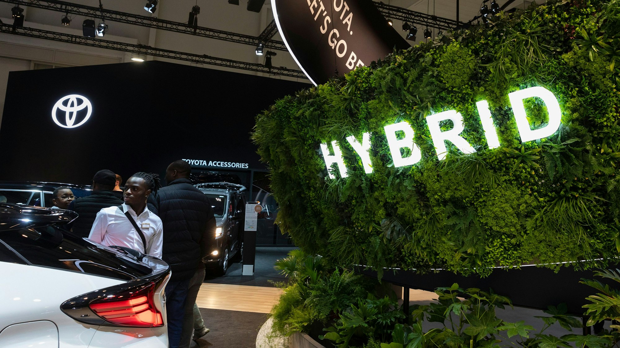 Abgebildet ist der Brüsseler Autosalon. Es sind Menschen und Autos sowie das Toyota-Logo zu sehen. Im Vordergrund ist eine Hecke mit einer Leuchtaufschrift „Hybrid“ zu sehen.