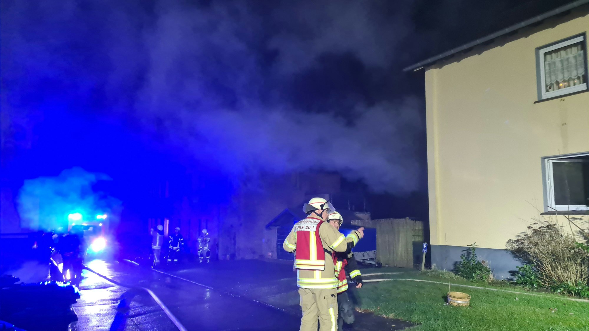 Feuerwehrleute stehen nachts im Scheinwerferlicht vor einem Wohnhaus, aus dem Brandrauch quillt.