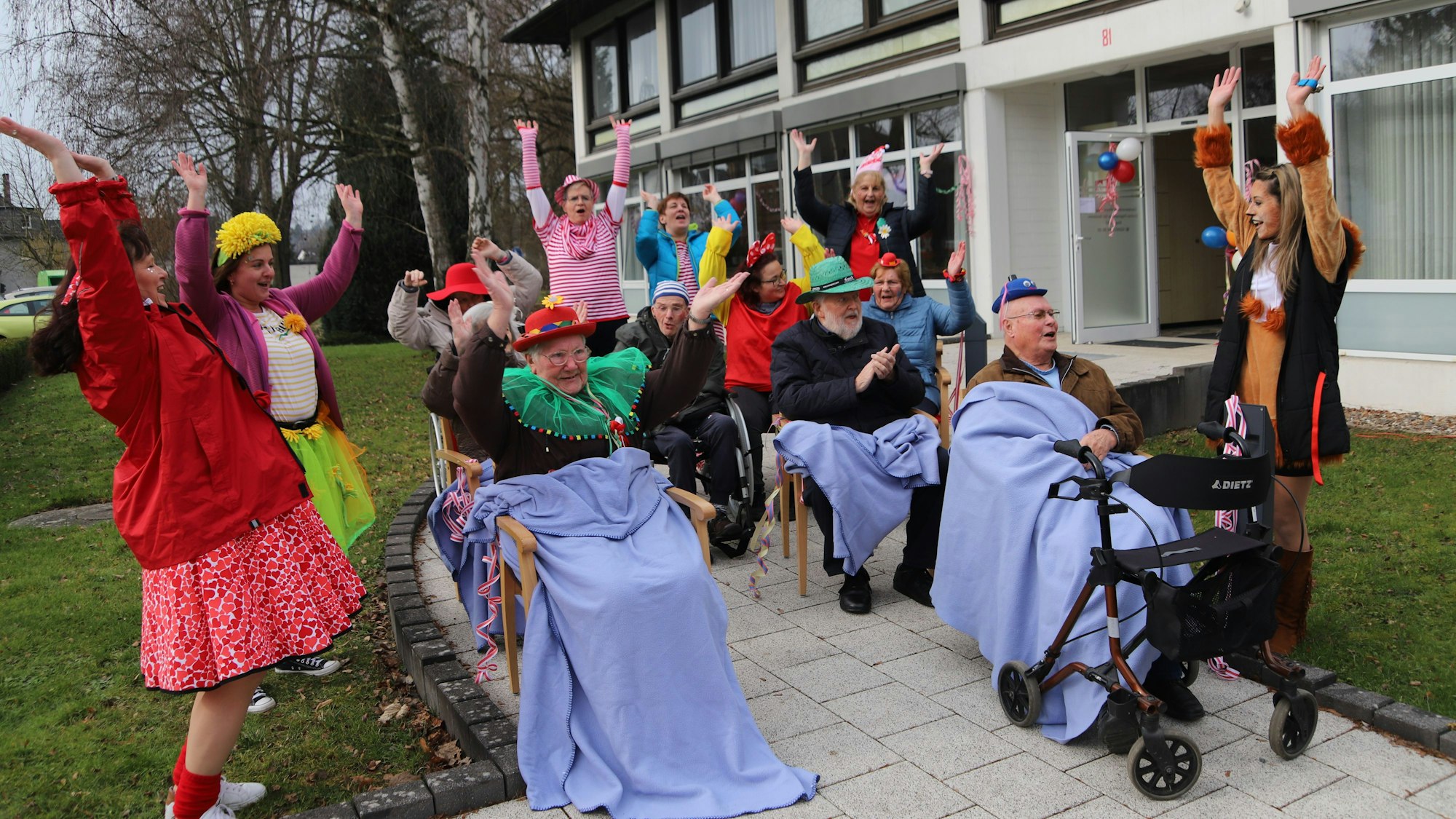 Kostümierte Senioren sitzen mit Wolldecken in Stühlen auf einem gepflasterten Weg, sie klatschen und haben die Arme in die Luft gehoben. Auch kostümierte Frauen neben und hinter ihnen reißen die Arme hoch.