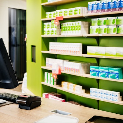 Das Bild zeigt eine Apotheke mit Kasse und einem Regal mit Medikamenten.