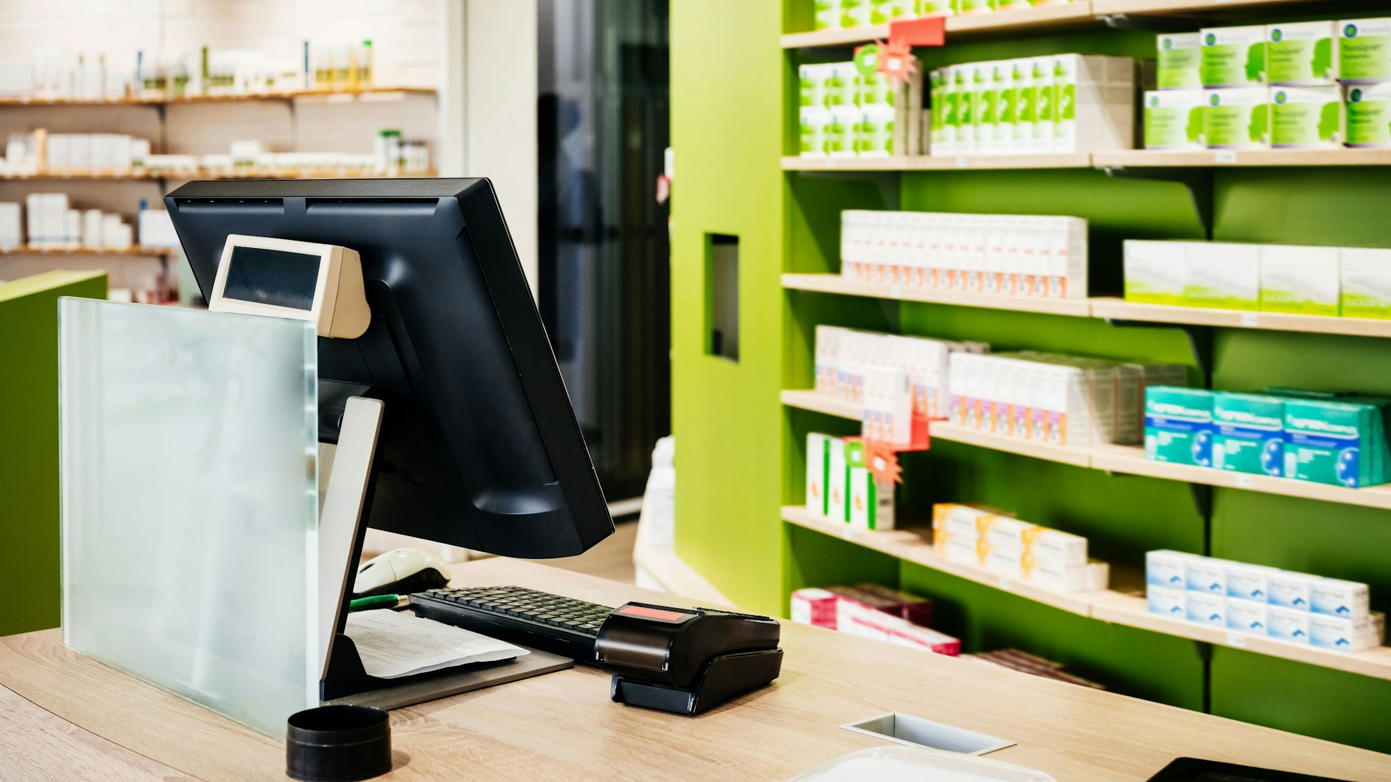 Das Bild zeigt eine Apotheke mit Kasse und einem Regal mit Medikamenten.