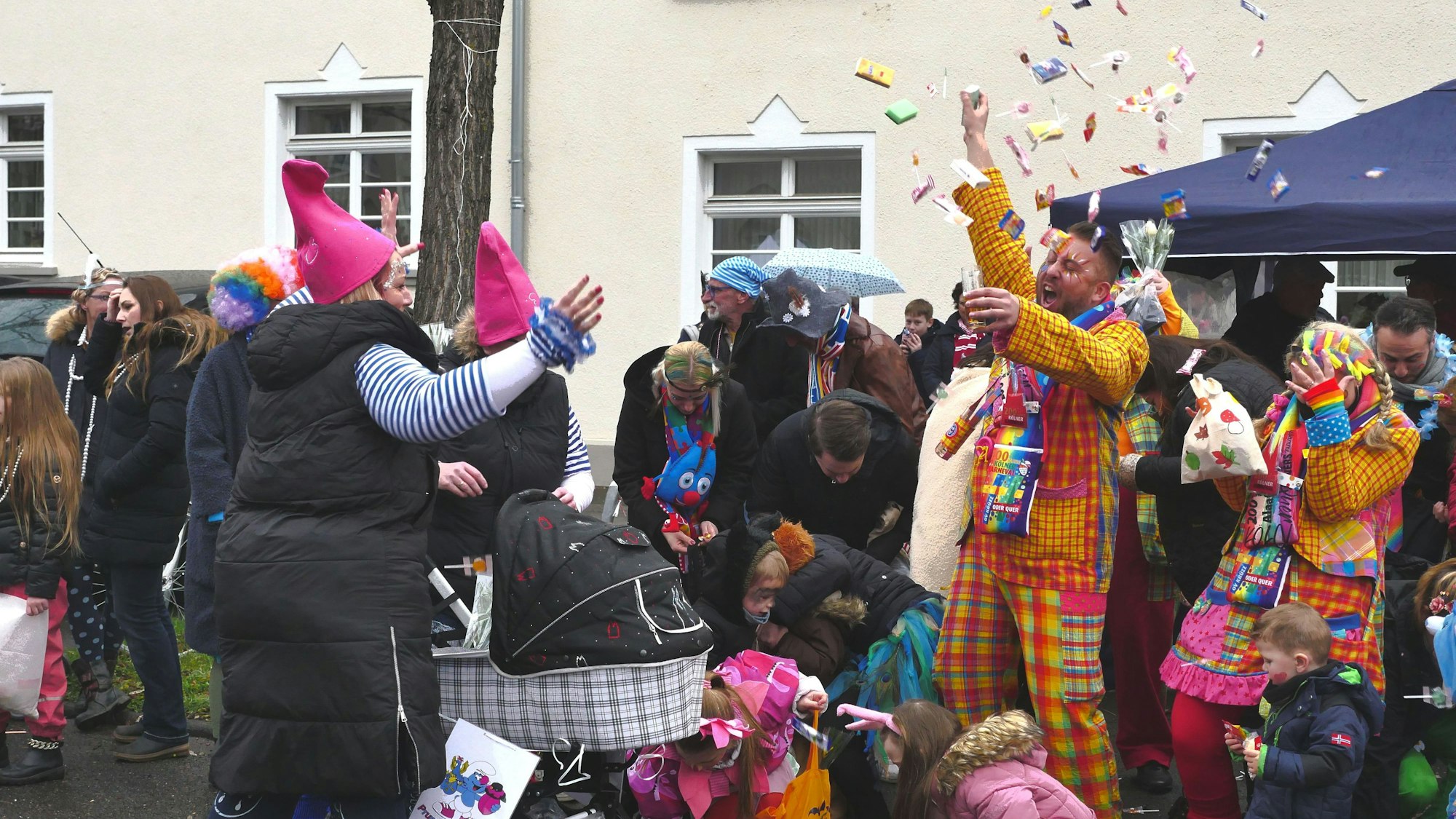 Ein Zugteilnehmer in buntem Outfit wirft Kamelle in die Luft. Kinder sammeln alles auf.