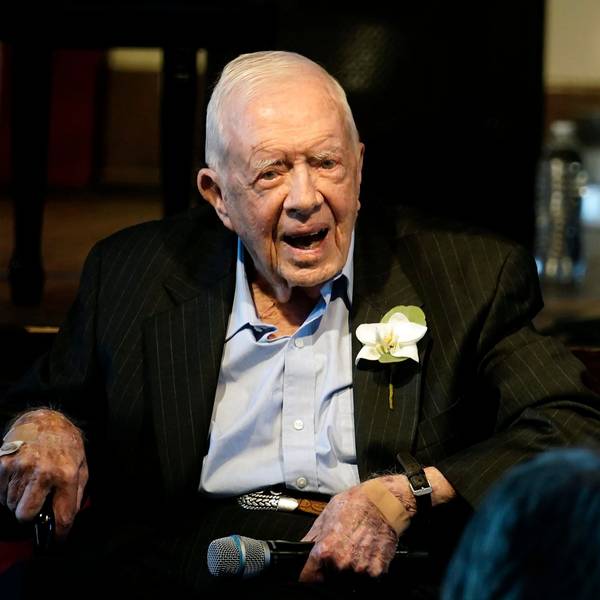 Ex-US-Präsident Jimmy Carter sitzt auf einem Stuhl und lächelt in die Kamera. Er trägt einen Anzug, in dessen Knopfloch eine Blume gesteckt ist.