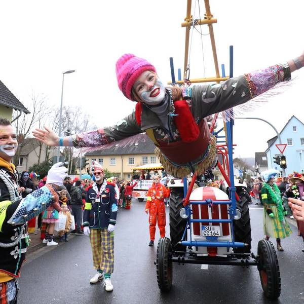 Eine bunte kostümierte Clownin schwebt als fliegender Clown an einer speziellen Konstruktion eines Traktors. Am Straßenrand stehen viele ebenfalls kostümierte Zugbesucher.