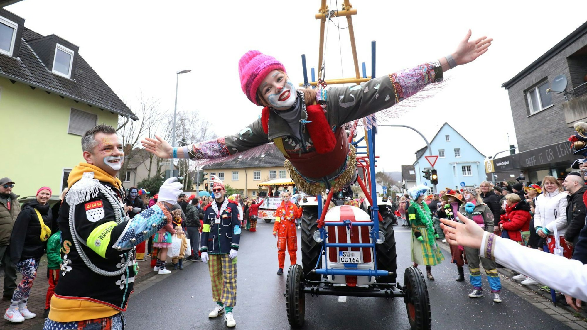Eine bunte kostümierte Clownin schwebt als fliegender Clown an einer speziellen Konstruktion eines Traktors. Am Straßenrand stehen viele ebenfalls kostümierte Zugbesucher.