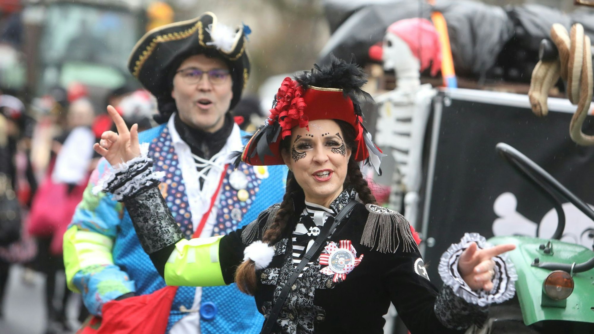 Eine Frau hat sich mit schwarzer Jacke und roter Mütze als Piratin verkleidet, am Karnevalswagen hinter ihr hängt ein Skelett mit roter Pappnase.