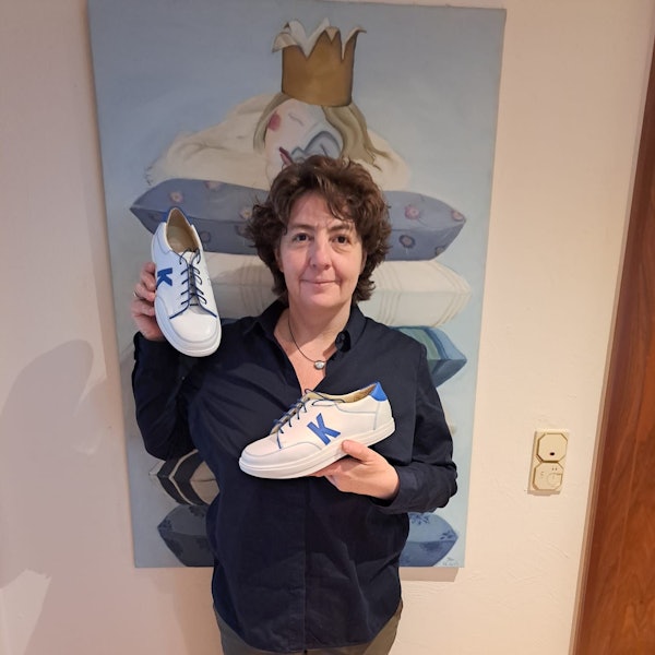 Beate Kandler steht vor einem blauen Bild und hält ihre Schuhe in die Kamera.