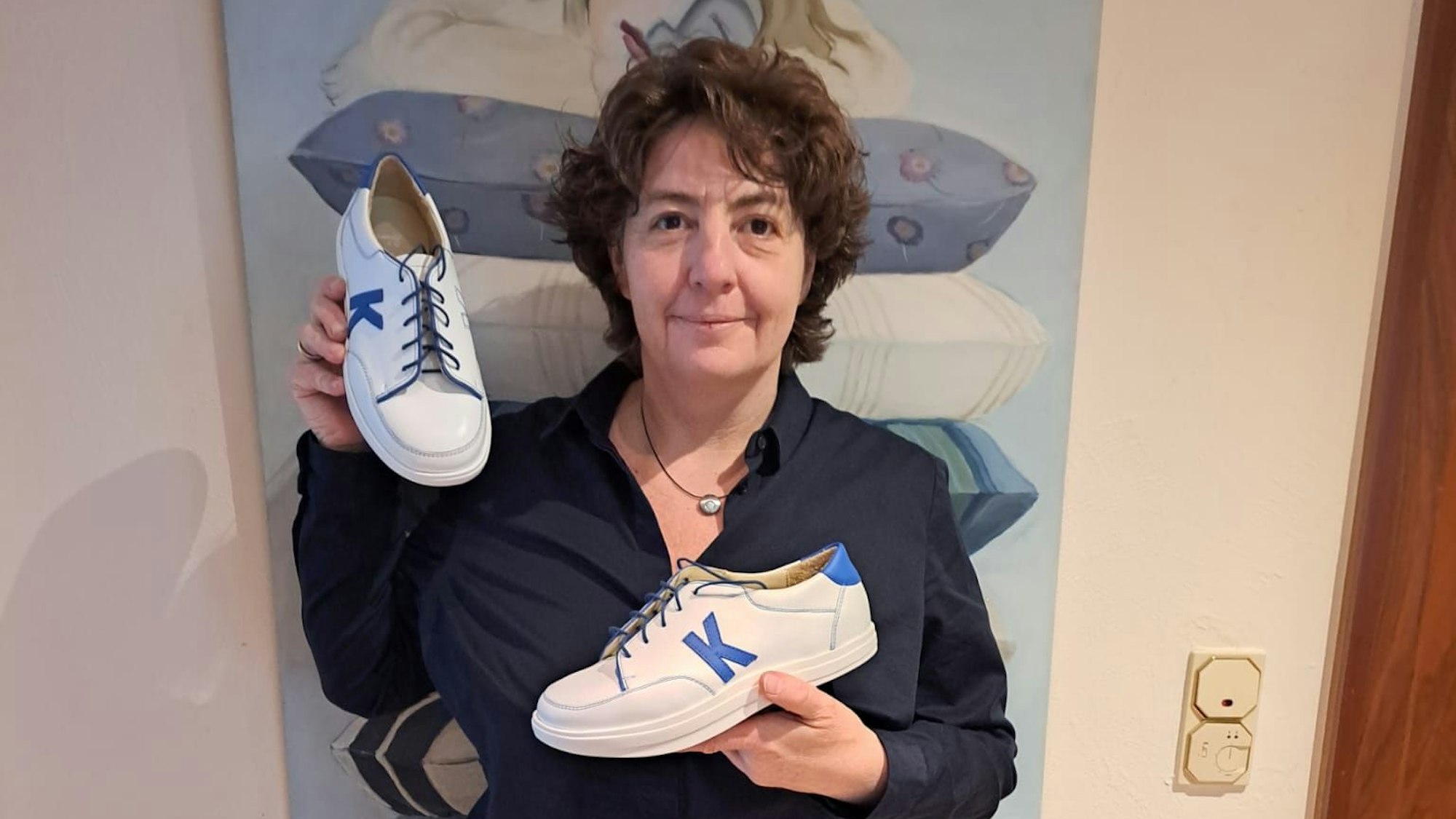 Beate Kandler steht vor einem blauen Bild und hält ihre Schuhe in die Kamera.