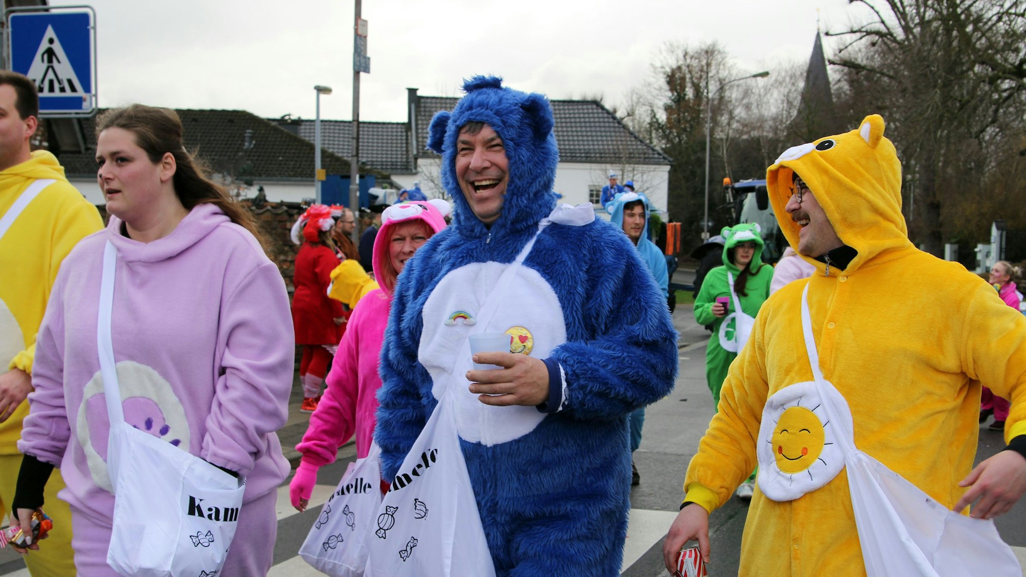 Als Glücksbärchen verkleidete Menschen gehen lachend über eine Straße.