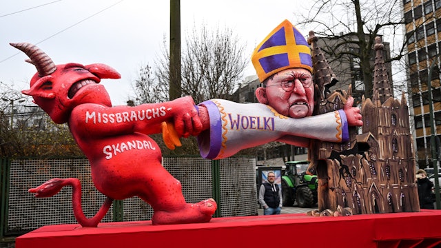 Ein Mottowagen beim Rosenmontagszug 2023 befasst sich mit dem Missbrauchsskandal in der katholischen Kirche. Die satirische Darstellung zeigt den Teufel, der am Kölner Kardinal Woelki zerrt. Dieser klammert sich an einem der beiden Türme des Kölner Doms fest, der aber bereits abzubrechen droht.