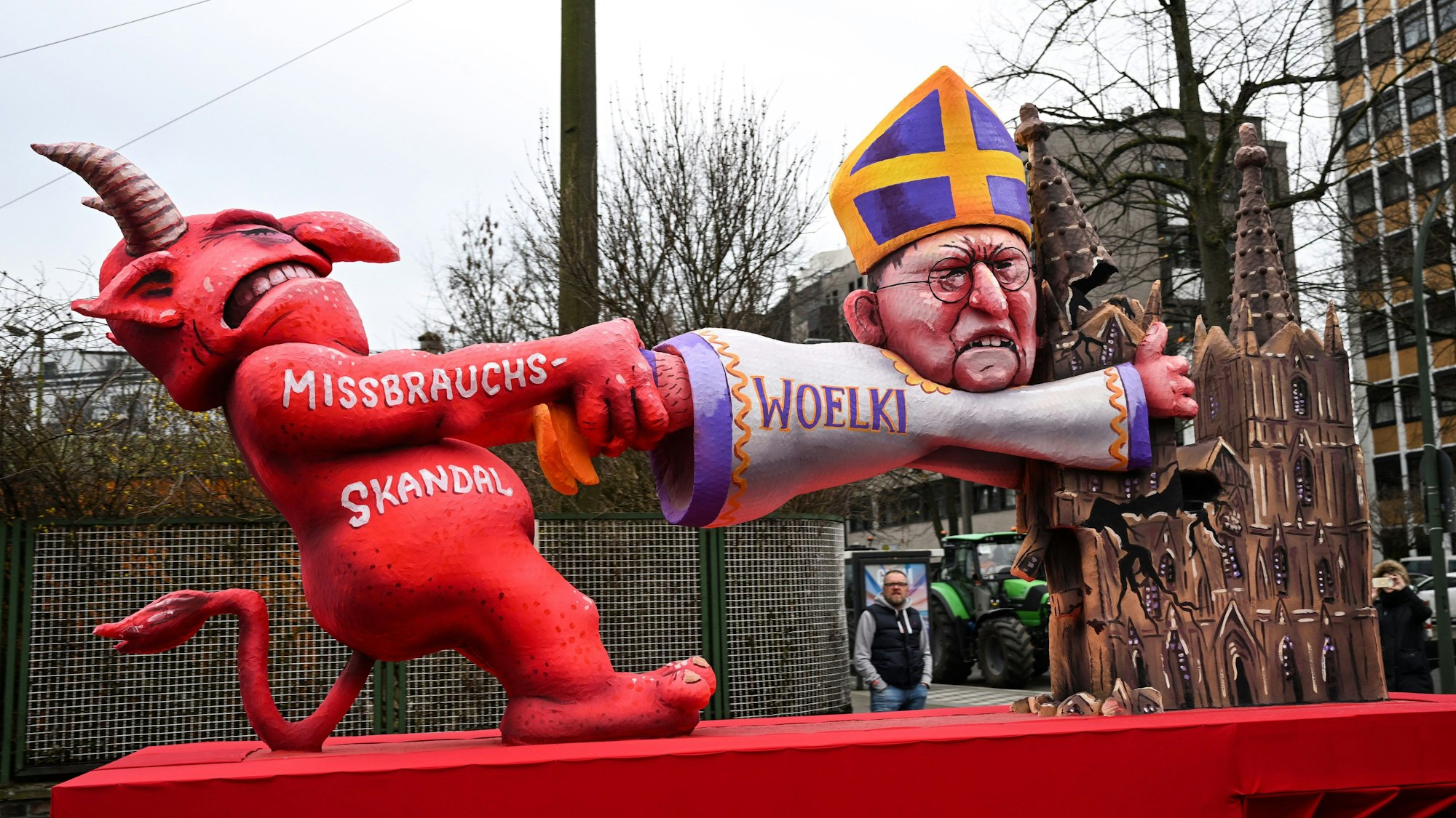 Ein Mottowagen beim Rosenmontagszug 2023 befasst sich mit dem Missbrauchsskandal in der katholischen Kirche. Die satirische Darstellung zeigt den Teufel, der am Kölner Kardinal Woelki zerrt. Dieser klammert sich an einem der beiden Türme des Kölner Doms fest, der aber bereits abzubrechen droht.