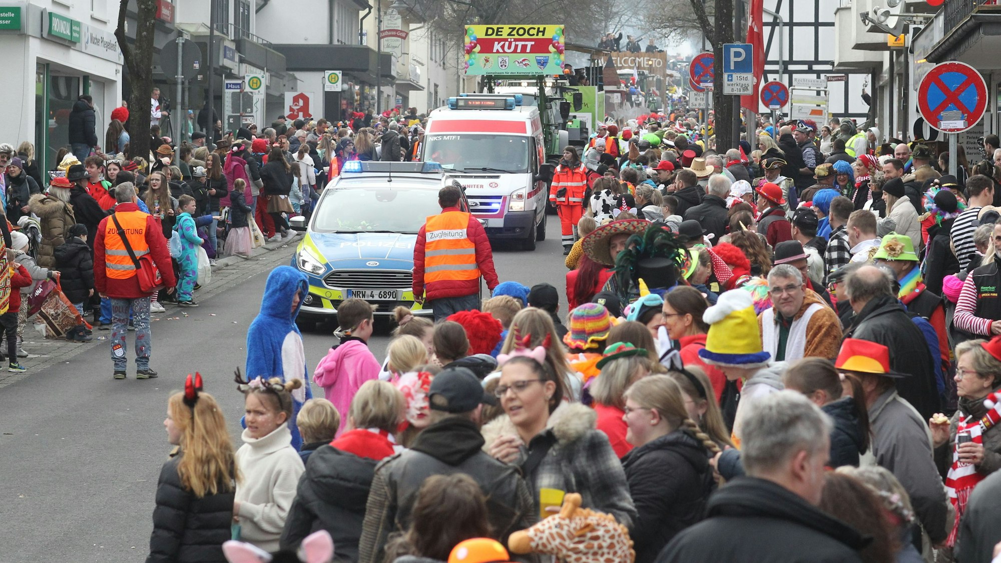 Menschen warten am Straßenrand auf einen Karnevalszug. Im Hintergrund ist ein Transparent zu sehen "De Zoch kütt" steht darauf, vorneweg fährt ein Polizeiauto.