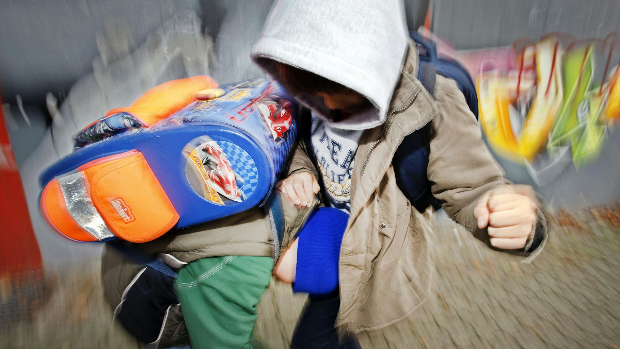 Zwei Schüler prügeln sich auf dem Schulhof. Ihre Gesichter sind nicht zu erkennen, der Hintergrund zeigt verschwommen eine Mauer mit Graffiti.