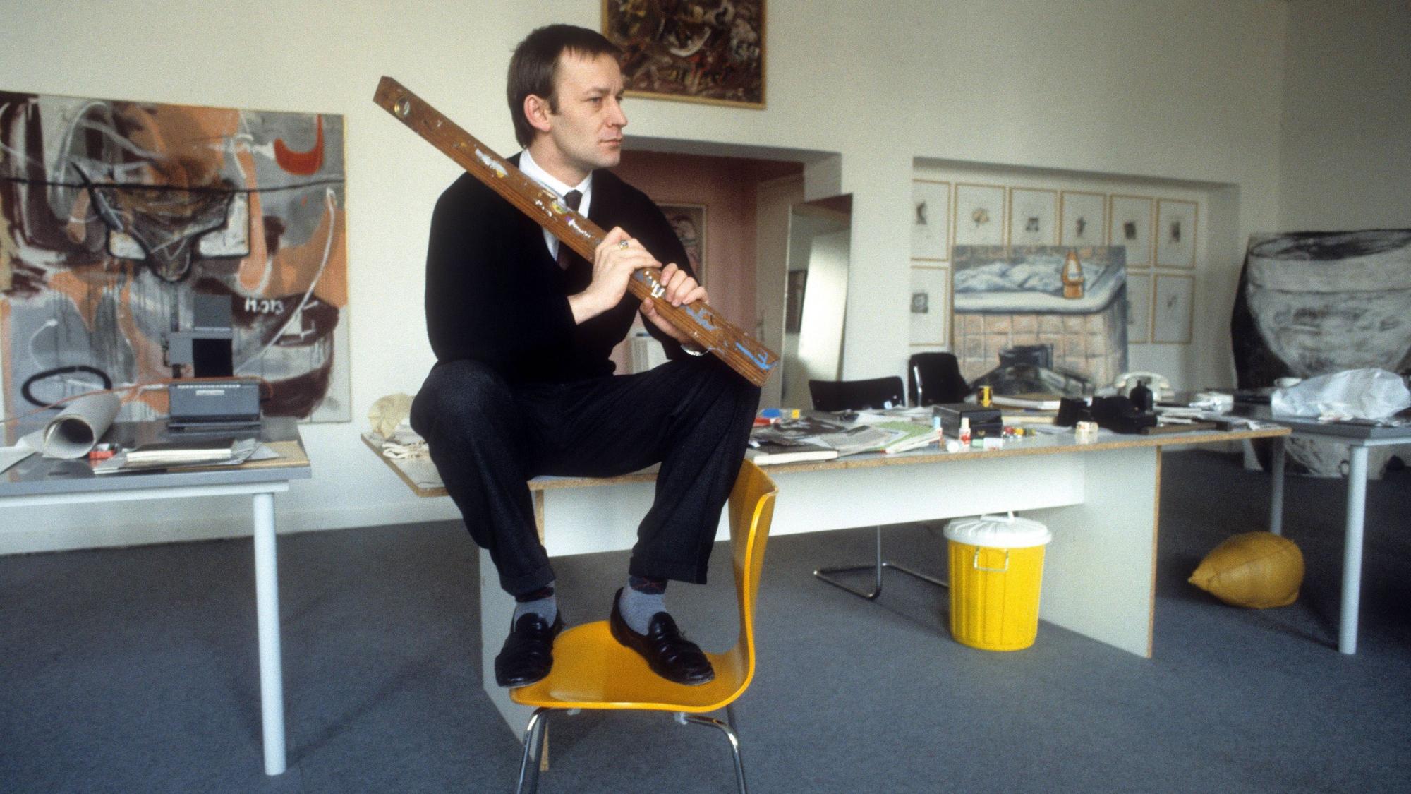 Martin Kippenberger sitzt auf einem Tisch, seine Füße stehen auf einem Stuhl.