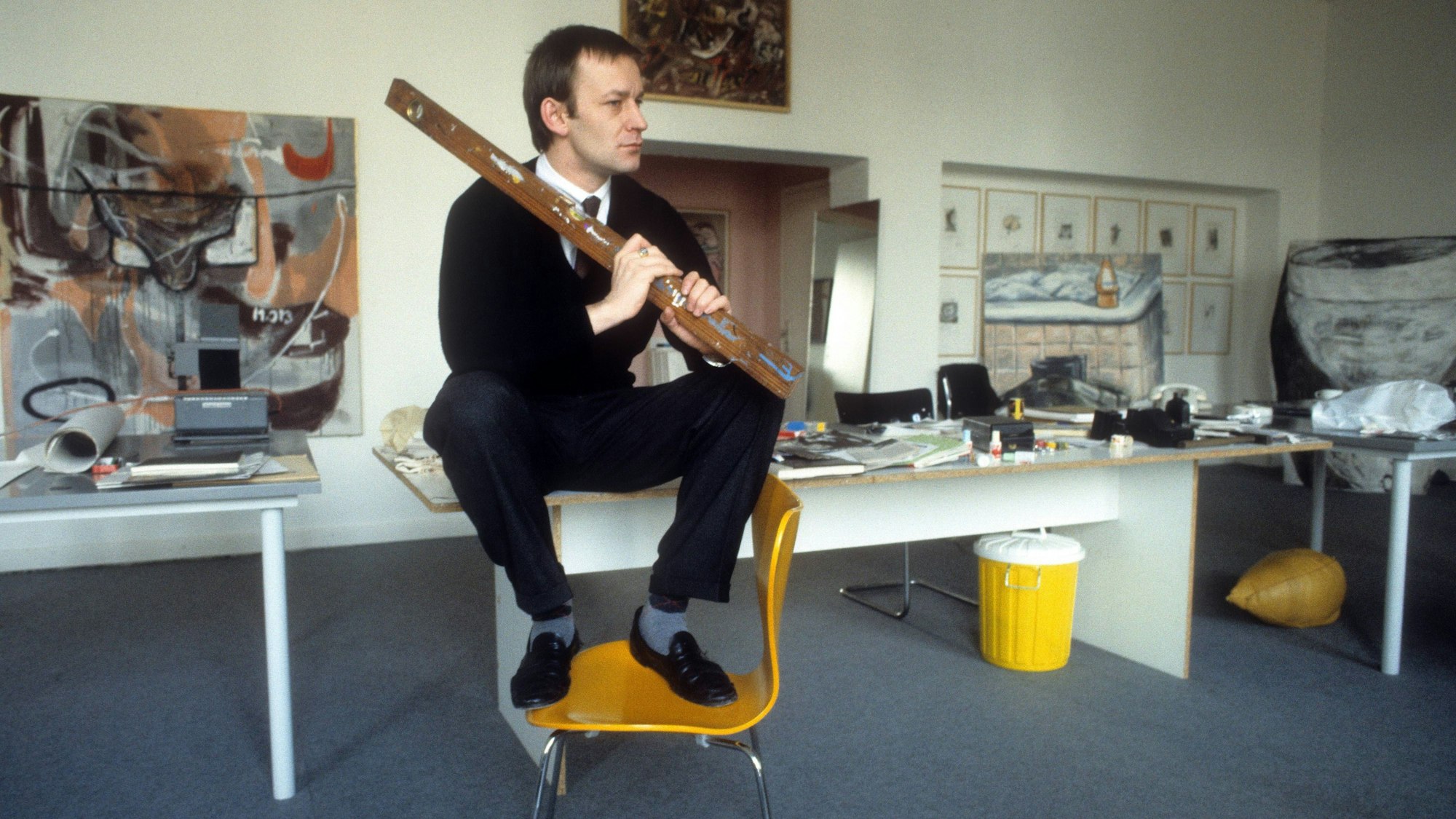 Martin Kippenberger sitzt auf einem Tisch, seine Füße stehen auf einem Stuhl.