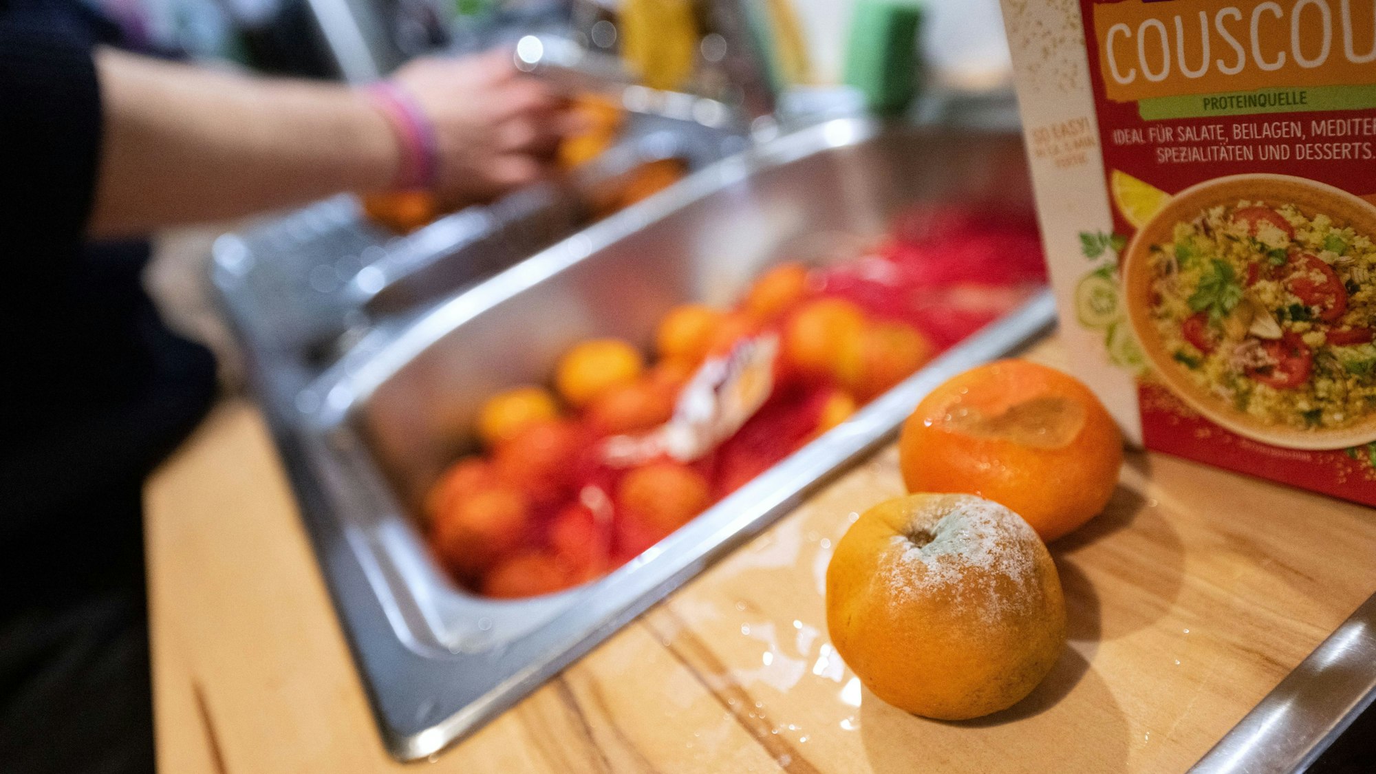 Einzelne schimmelige Mandarinen liegen aussortiert auf einer Küchenarbeitsplatte, während eine Frau andere unverdorbene Mandarinen wäscht.