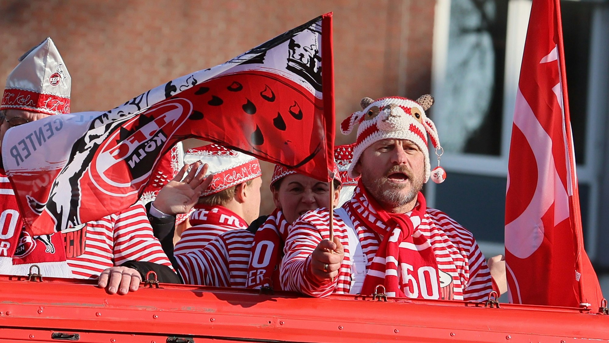 FC-Trainer Steffen Baumgart schwenkt auf dem Kölner Rosenmontagszug auf einem Wagen eine Flagge des 1. FC Köln. Auf dem Kopf trägt er eine gehäkelte Geißbock-Mütze.