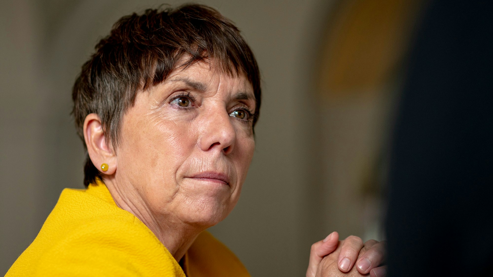 Die evangelische Theologin Margot Käßmann wendet während eines Interviews ihr Gesicht dem unsichtbaren Interviewer zu. Sie trägt einen gelben Blazer, hat die Arme aufgestützt und die Hände gefaltet.