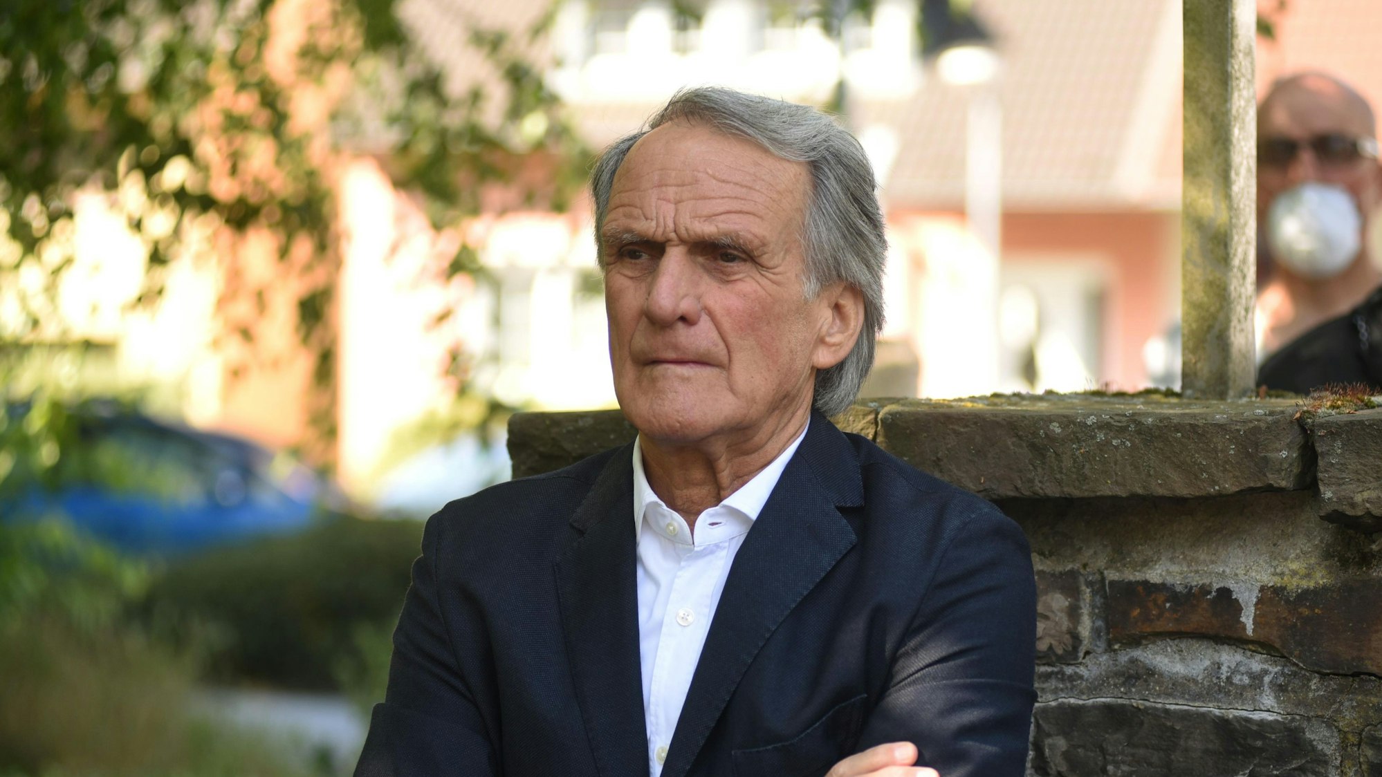 Wolfgang Overath mit grauen, längeren Haaren steht vor einer Mauer und hat die Arme vor der Brust überkreuzt.