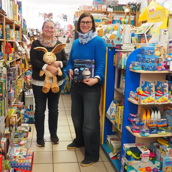 Zwei Frauen stehen mit Spielzeug in der Hand zwischen vollen bunten Regalen in ihrem Spielwarengeschäft.