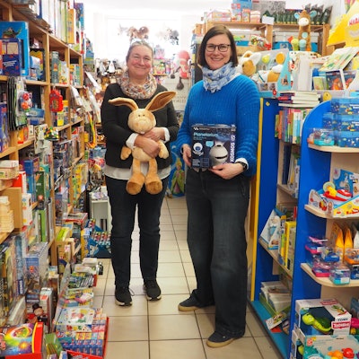 Zwei Frauen stehen mit Spielzeug in der Hand zwischen vollen bunten Regalen in ihrem Spielwarengeschäft.