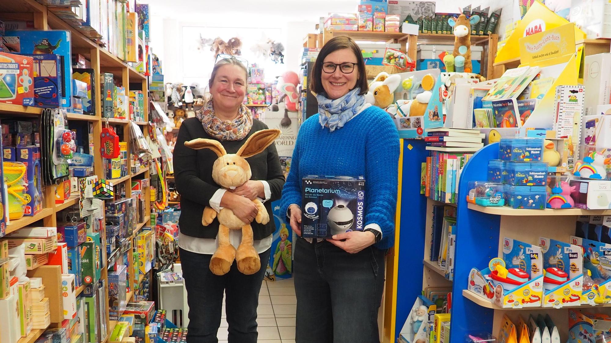 Zwei Frauen stehen mit Spielzeug in der Hand zwischen vollen bunten Regalen in ihrem Spielwarengeschäft.