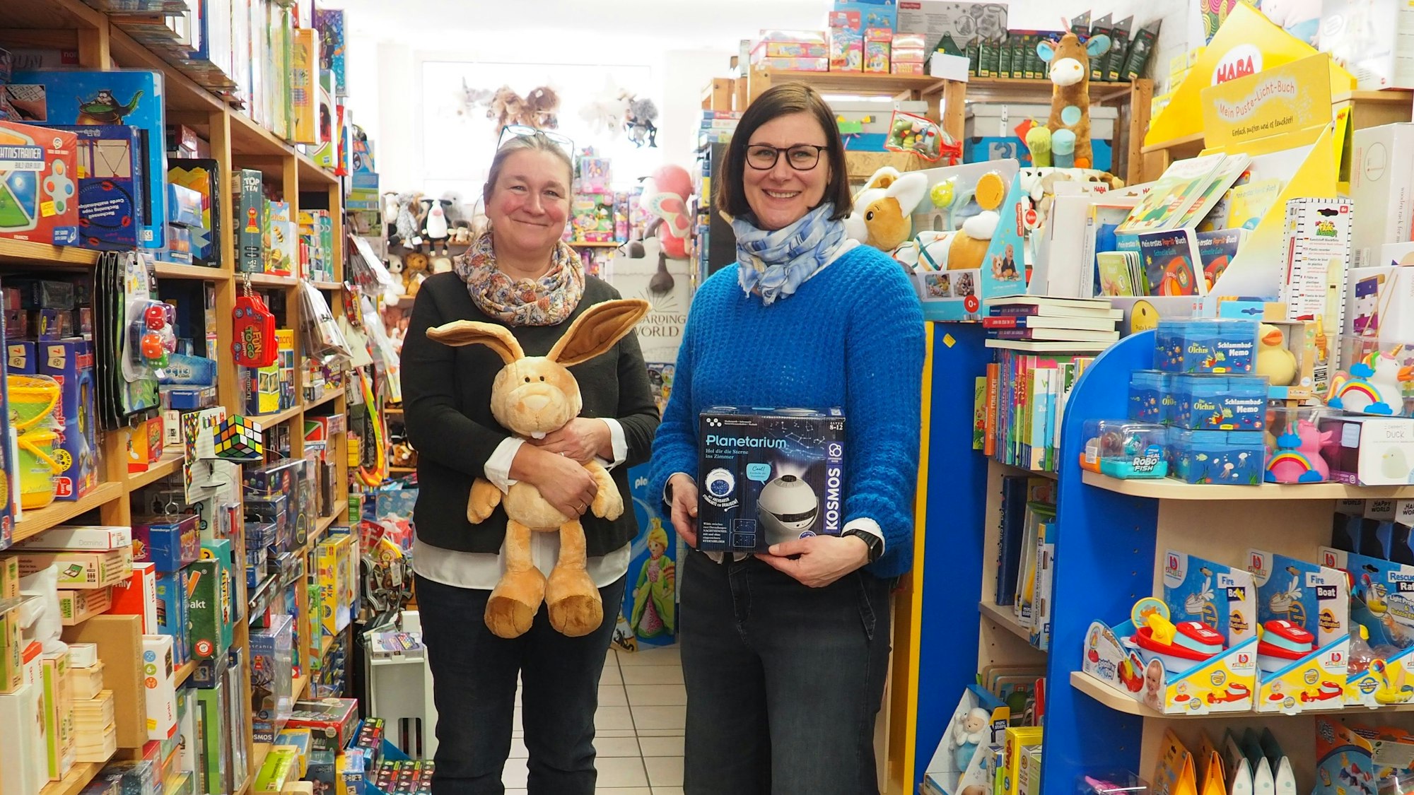 Zwei Frauen stehen mit Spielzeug in der Hand zwischen vollen bunten Regalen in ihrem Spielwarengeschäft.
