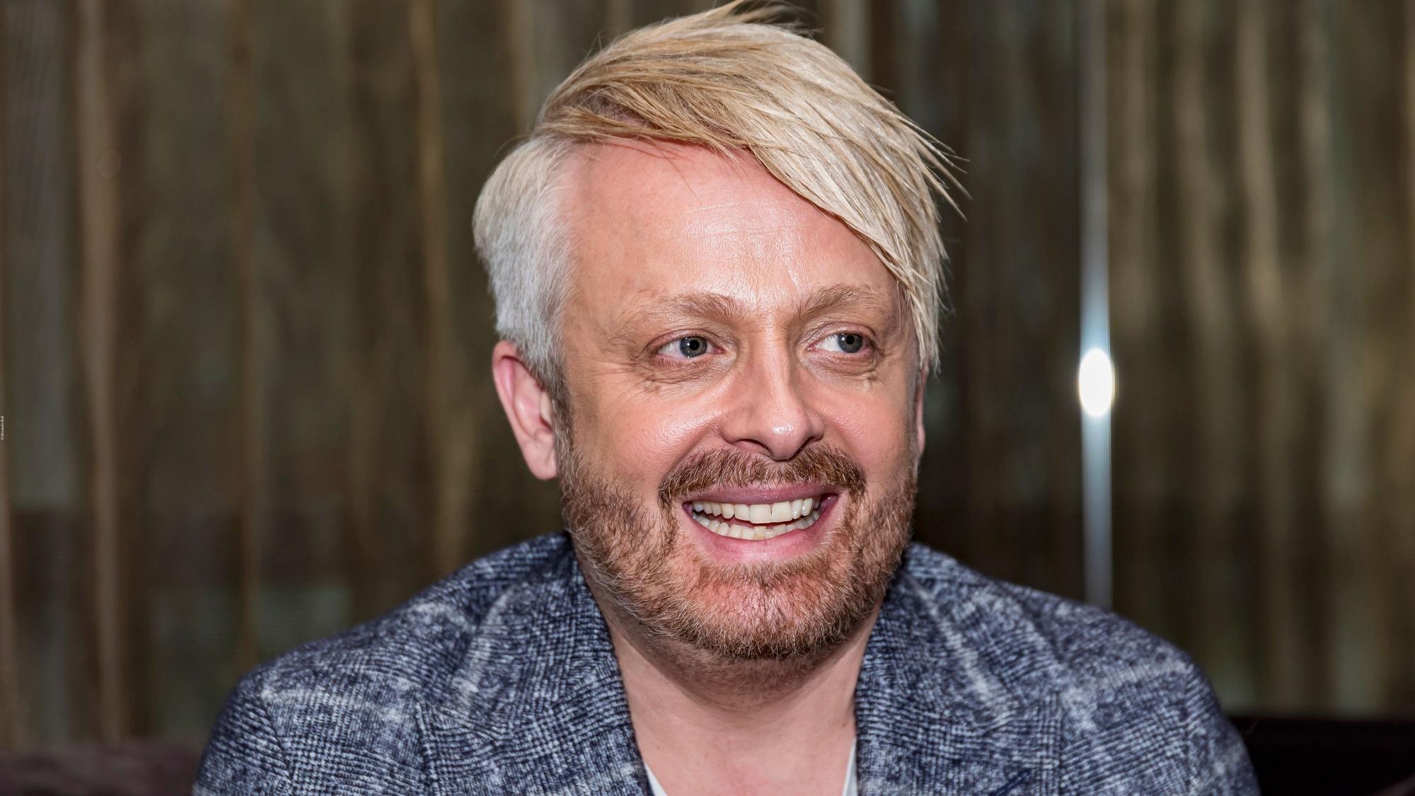 Ross Antony hat ein blau-weiß kariertes Sacko an und lacht an der Kamera vorbei.