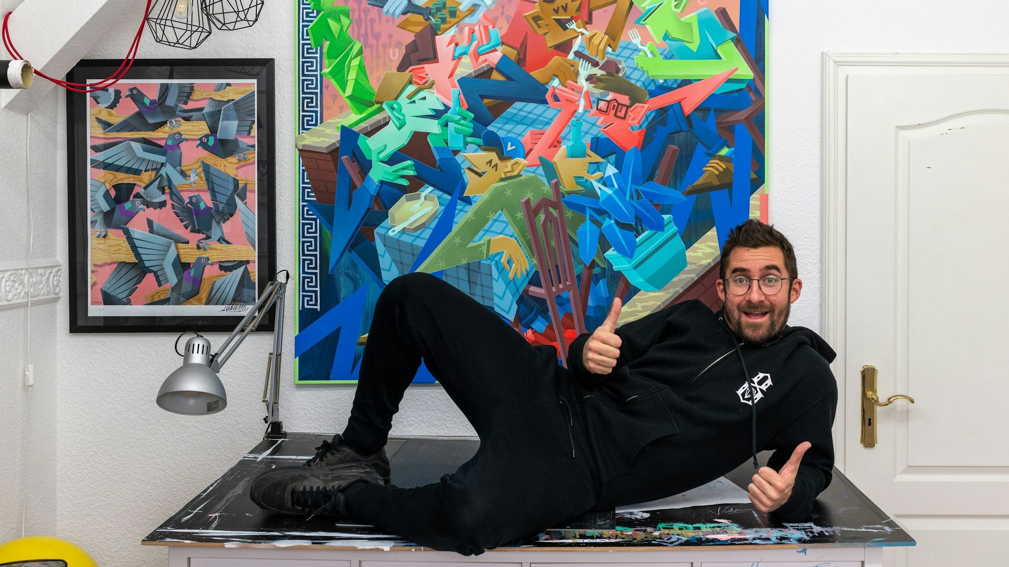 Ein Mann liegt auf einer Kommode und streckt beide Daumen in die Höhe. Hinter ihm ist ein buntes Graffito zu sehen.