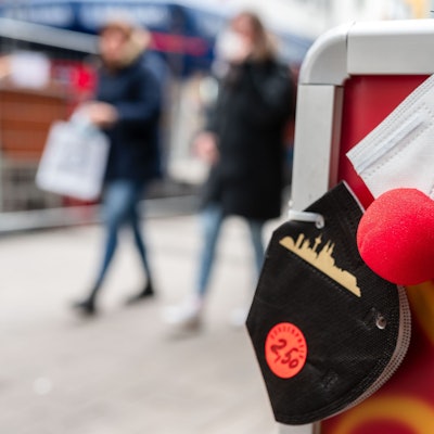 Eine FFP2-Maske mit einer roten Clownsnase hängt an einem Aufsteller vor einem Geschäft.