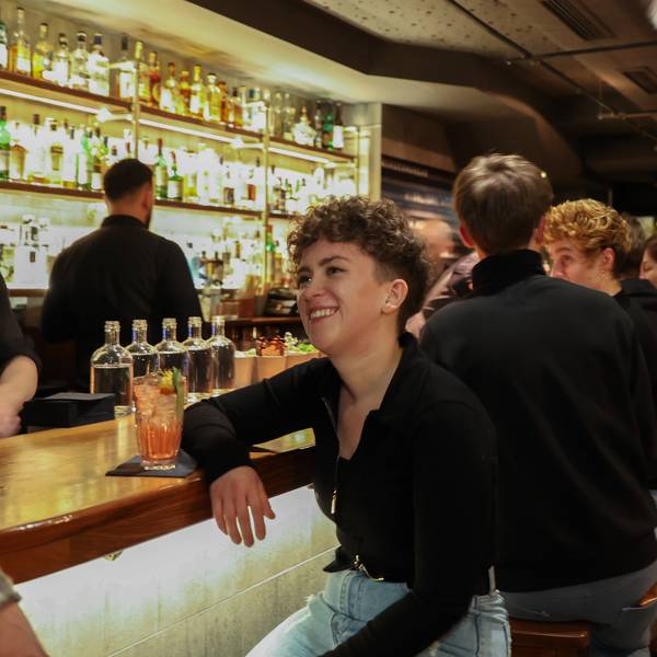 Eine junge Frau sitzt an der Bar. Vor ihr steht ein Drink, hinter ihr sitzen weitere Besucher.