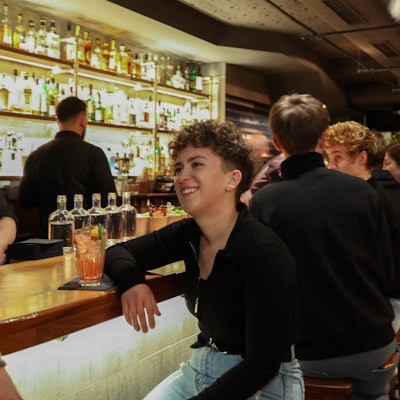 Eine junge Frau sitzt an der Bar. Vor ihr steht ein Drink, hinter ihr sitzen weitere Besucher.