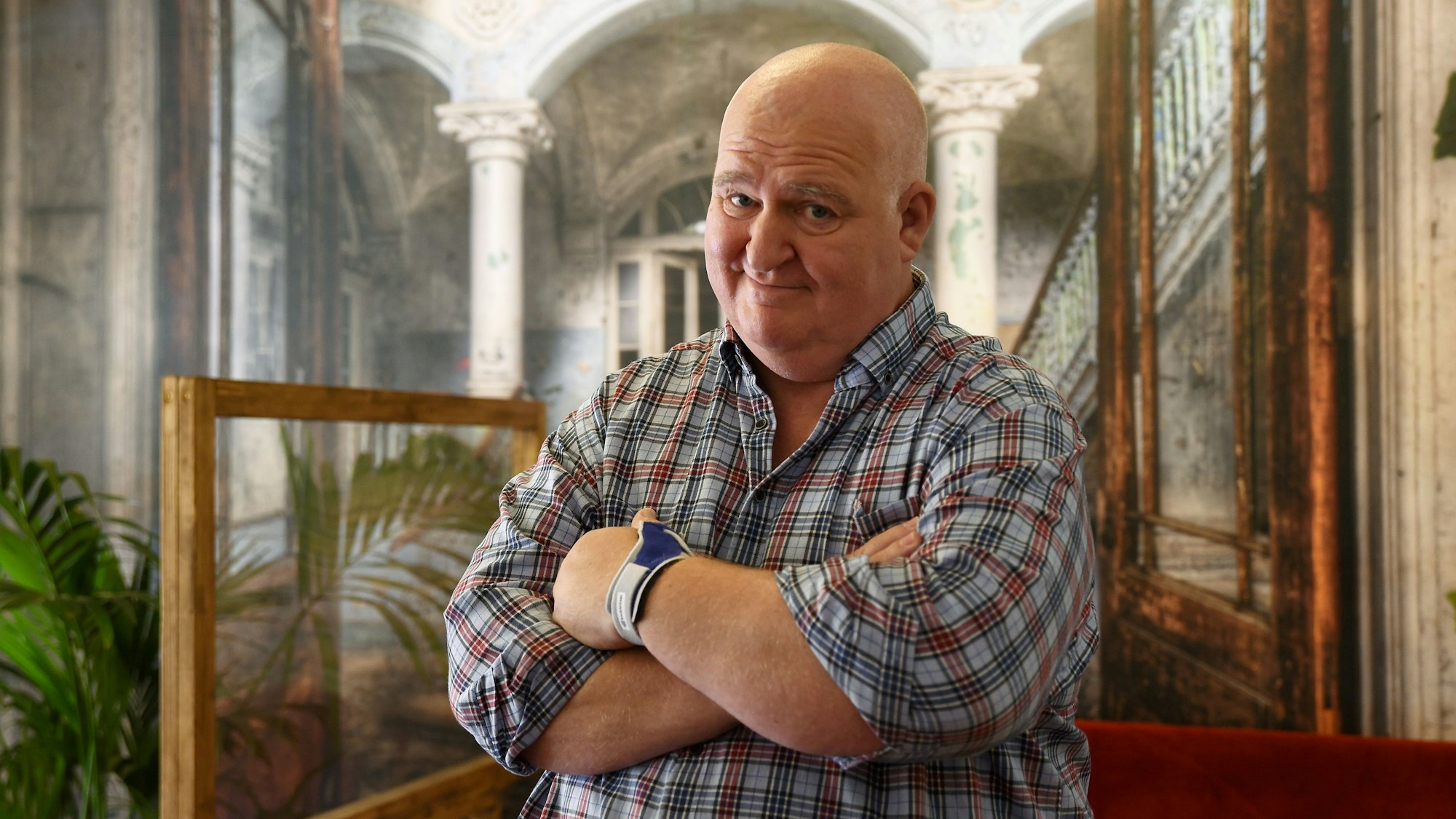Markus Maria Profitlich steht in einem Karo-Hemd mit den Armen vor der Brust verschränkt vor einer rustikalen Kulisse.