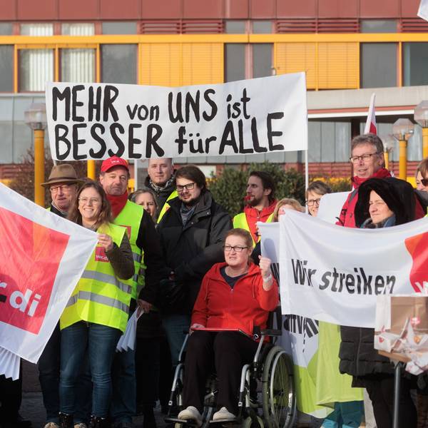 Bei einem Streik halten mehrere Personen, bekleidet mit gelben Warnwesten, Schilder und Banner in die Höhe. Darauf steht: „Wir streiken“, „Verdi“ und „Mehr von uns ist besser für alle“.