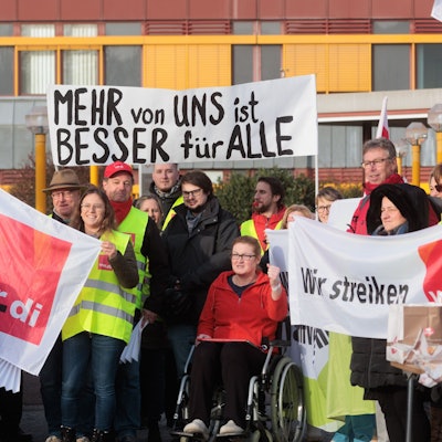 Bei einem Streik halten mehrere Personen, bekleidet mit gelben Warnwesten, Schilder und Banner in die Höhe. Darauf steht: „Wir streiken“, „Verdi“ und „Mehr von uns ist besser für alle“.