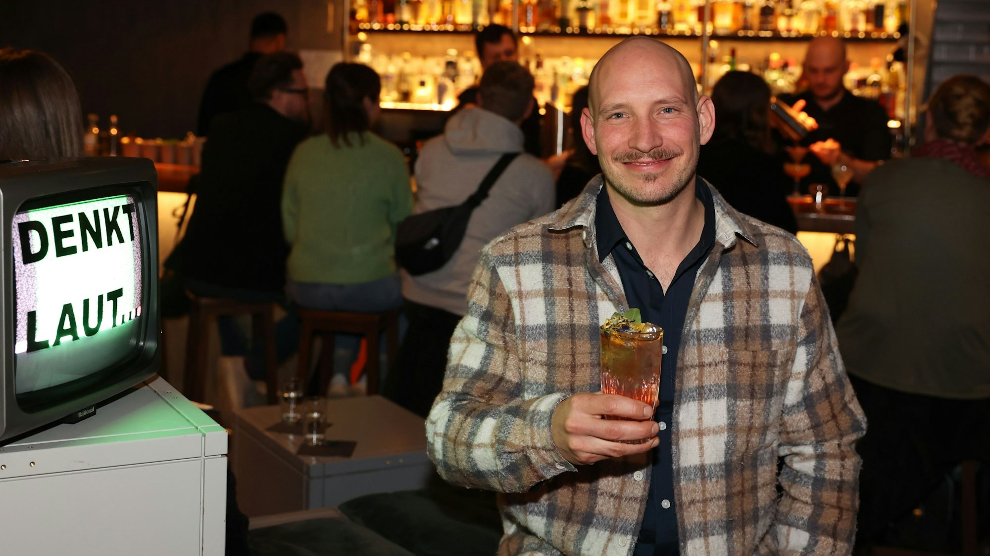 Felix Engels sitzt im Suderman. In der Hand hält er einen Drink. im Hntergrund ist die Bar zu sehen.