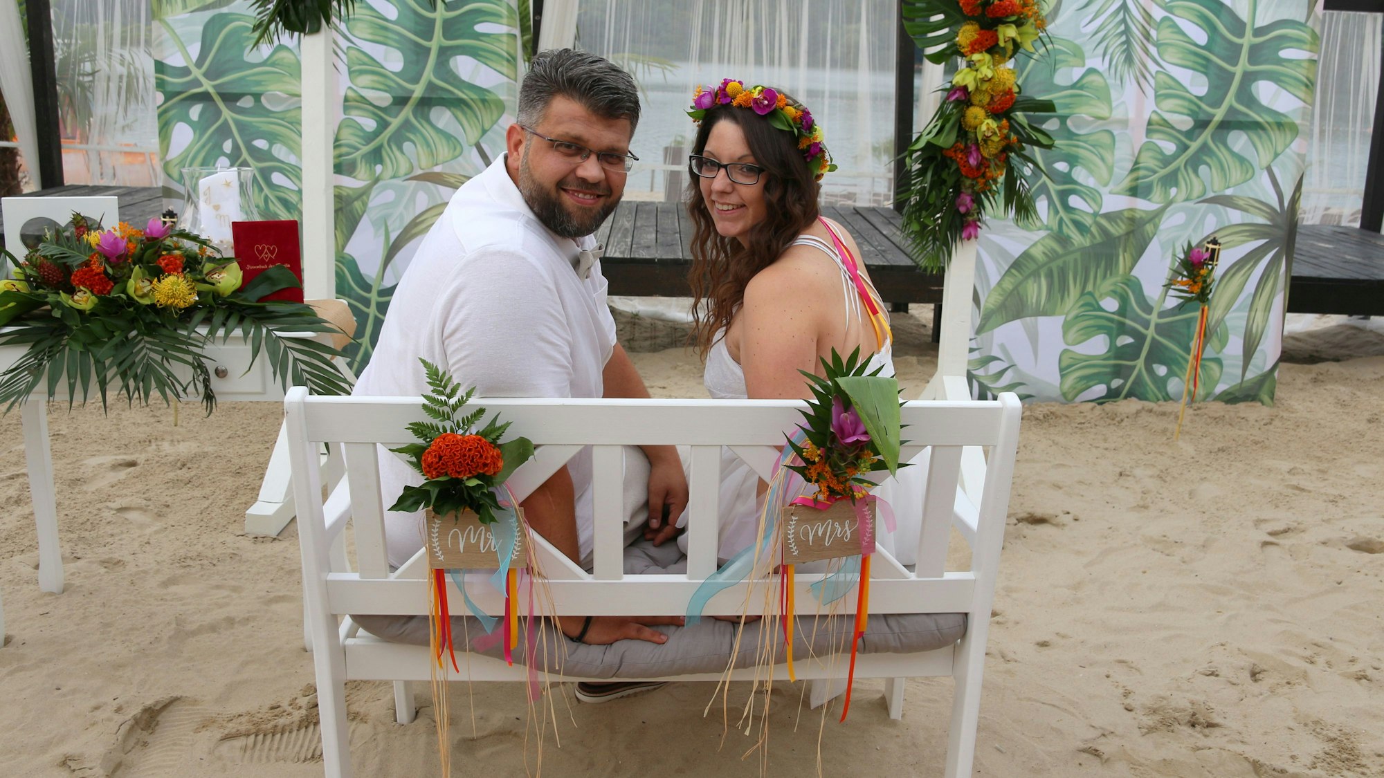 Es ist das Hochzeitsspaar auf einer Bank zu sehen, wie es sich umdreht und in die Kamera guckt.