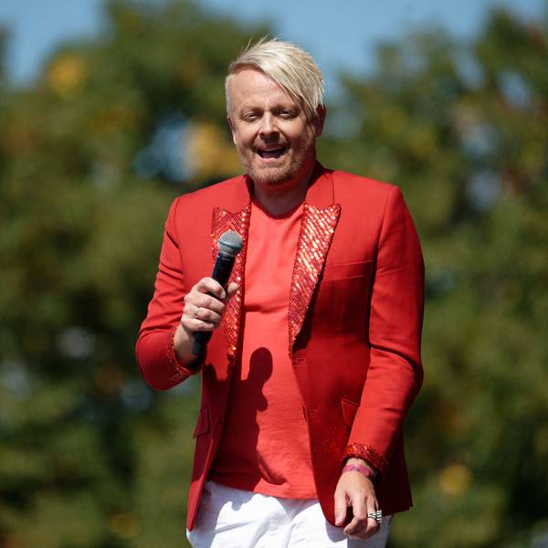 Ross Antony trägt ein rotes Sakko mit Glitzer, er hat ein Mikro in der Hand. Im Hintergrund sind Laubbäume zu erkennen.