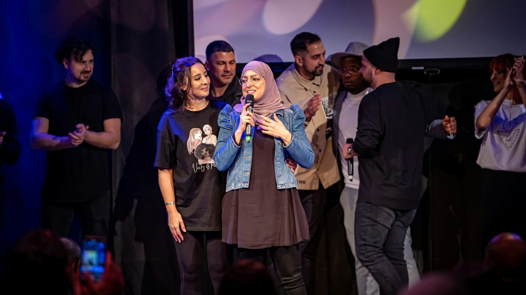 Die Veranstalterinnen stehen auf der Bühne, Asmaa El Idrissi hält ein Mikrofon in der Hand. Hinter ihnen stehen weitere Künstler, die während des Events auftraten.