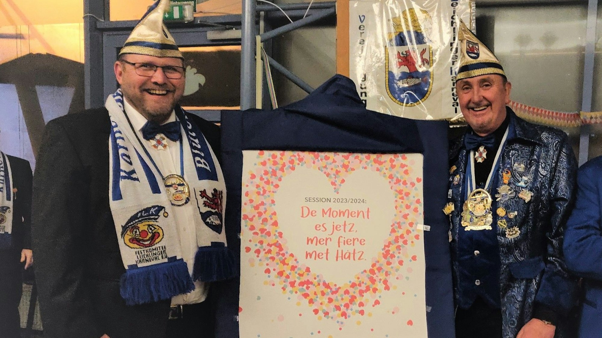 Zwei vom designierten Leichlingen Dreigestirn: Jungfrau Lars „Larissa“ Stenzel (l.) und Prinz Volkmar Blank stehen neben einer Tafel mit ihrem neuen Sessionsmotto „De Moment es jetz. Mer fiere met Hätz“