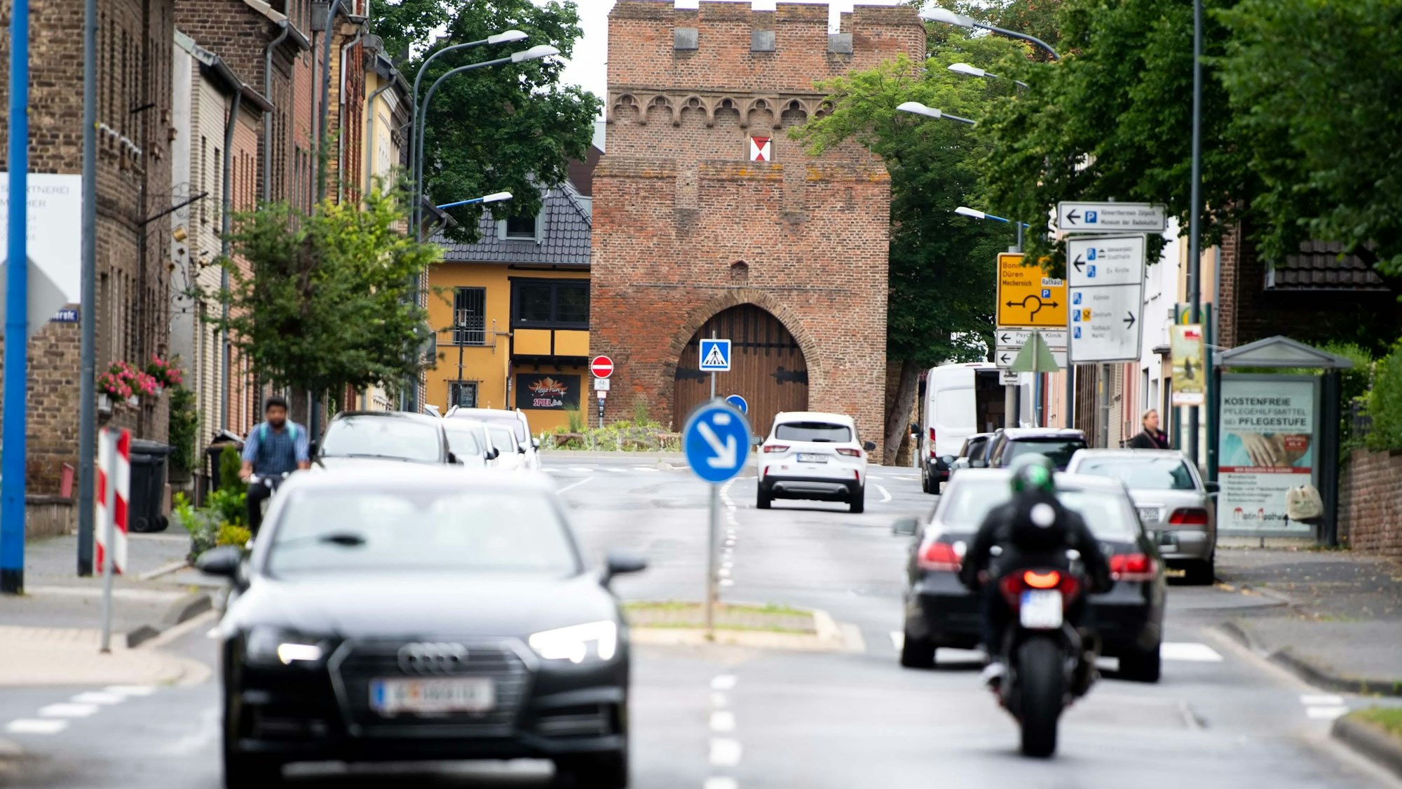 Das Bild zeigt einen Blick auf die Römerallee in Zülpich. Sie wird von mehreren Autos, einem Motorrad und einem Radfahrer befahren.