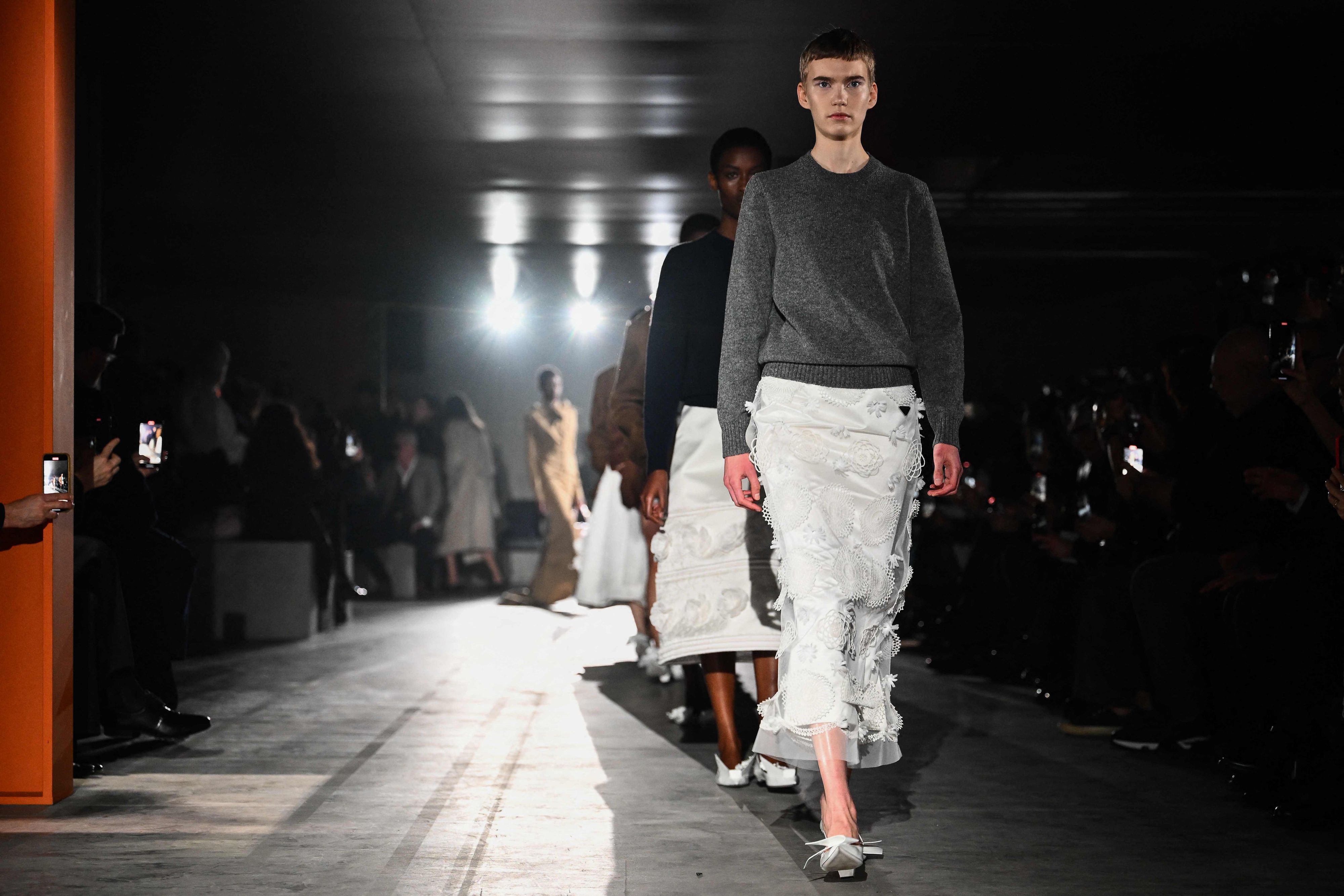 Models präsentieren Kreationen von Prada auf dem Laufsteg. Das erste Model trägt einen weißen, verschnörkelten Rock und einen schlichten grauen Pullover.