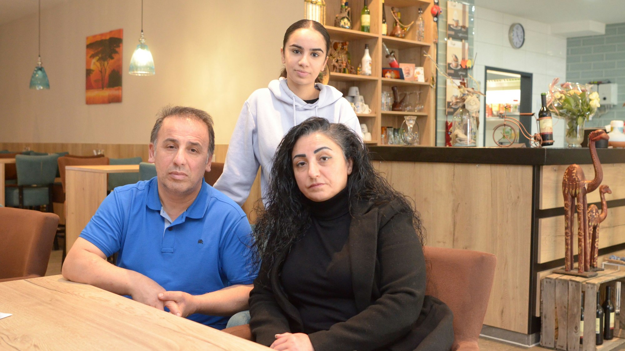 Die drei Personen sind an einem Tisch zu sehen, die Eltern sitzen, die Tochter steht hinter ihnen.