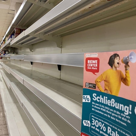 Man sieht ein leeres Regal, rechts ein Rewe Plakat, Thema Schließung.