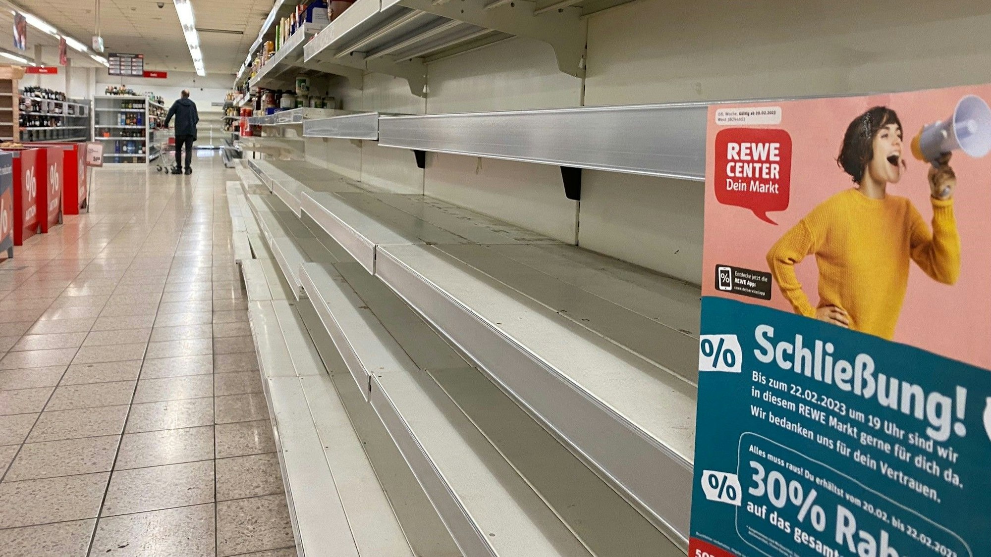 Man sieht ein leeres Regal, rechts ein Rewe Plakat, Thema Schließung.