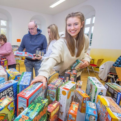 Eine Frau steht vor einem Tisch, auf dem Kartons mit Gesellschaftsspielen gestapelt sind. Sie greift nach einem Karton.
