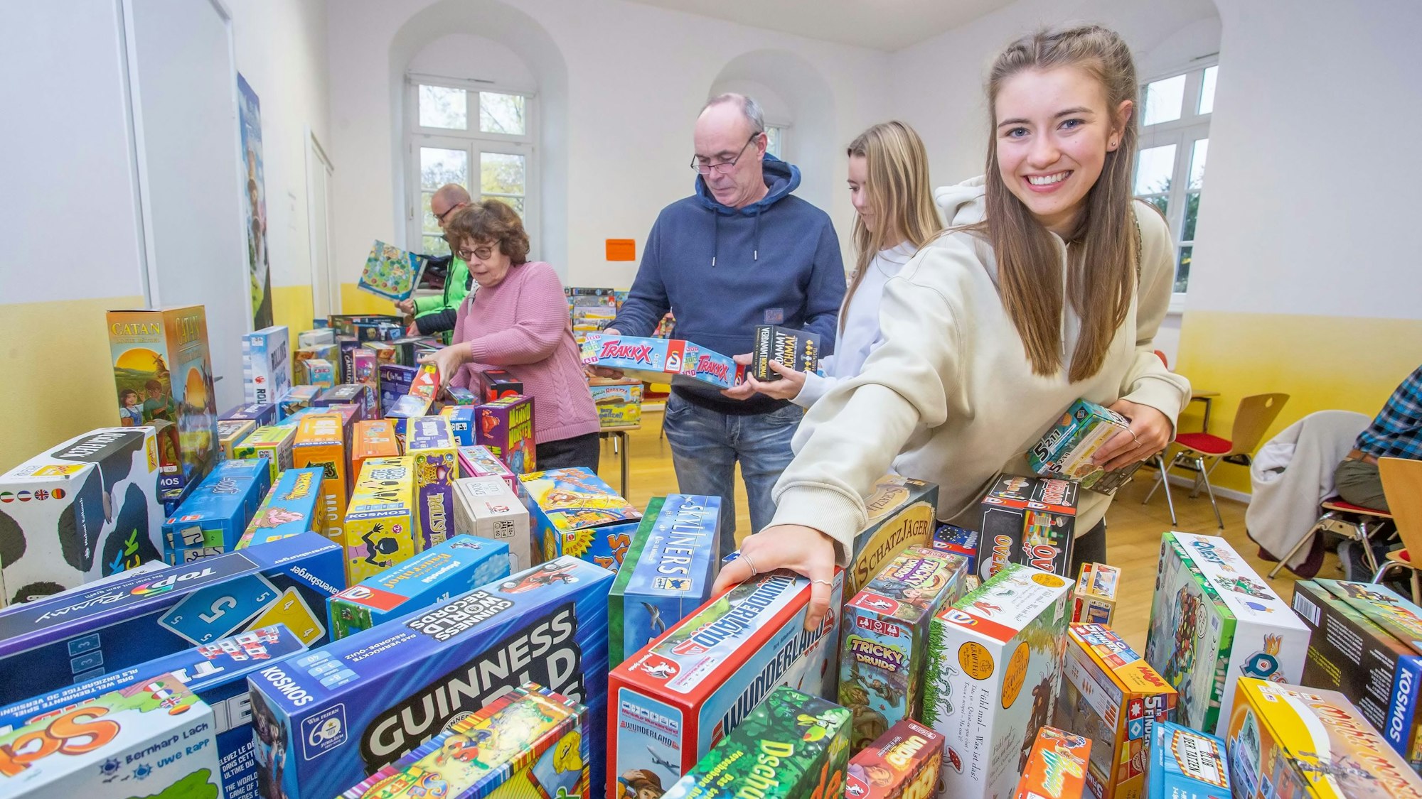 Eine Frau steht vor einem Tisch, auf dem Kartons mit Gesellschaftsspielen gestapelt sind. Sie greift nach einem Karton.